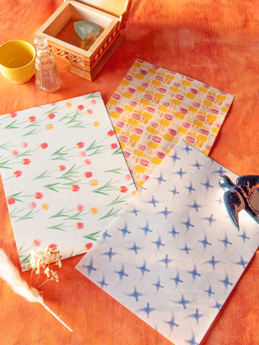 オレンジ色の布の上に3枚のデザインペーパーが並べられている。 左はチューリップ柄、中央は黄色とピンクの四角柄、右は青いつばめ柄。 周囲には木箱、黄色のカップ、ガラス瓶、青い鳥の置物、小さなドライフラワーが飾られている。
