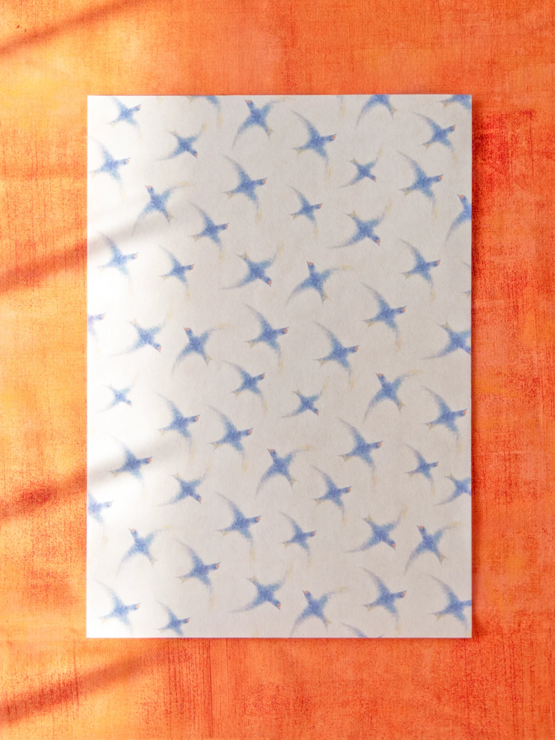 オレンジ色の布の上に置かれたデザインペーパー。青いツバメが舞うように飛んでいる柄。
